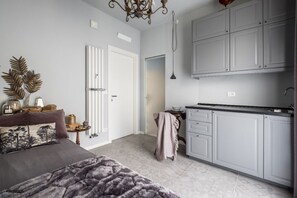 A modern bedroom with a large bed, a bedside table with decorative items, and a view into the adjacent kitchen with grey cabinets and a hanging light fixture. - Fico (Bologna)