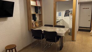 A dining area with a table and chairs, a bookshelf, a television, and a door leading to a kitchen.
