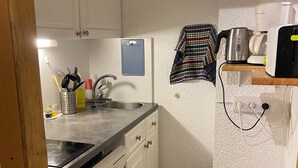 A compact kitchen with white cabinets, a stainless steel sink, a small trash bin, and a shelf with a kettle and a coffee maker.