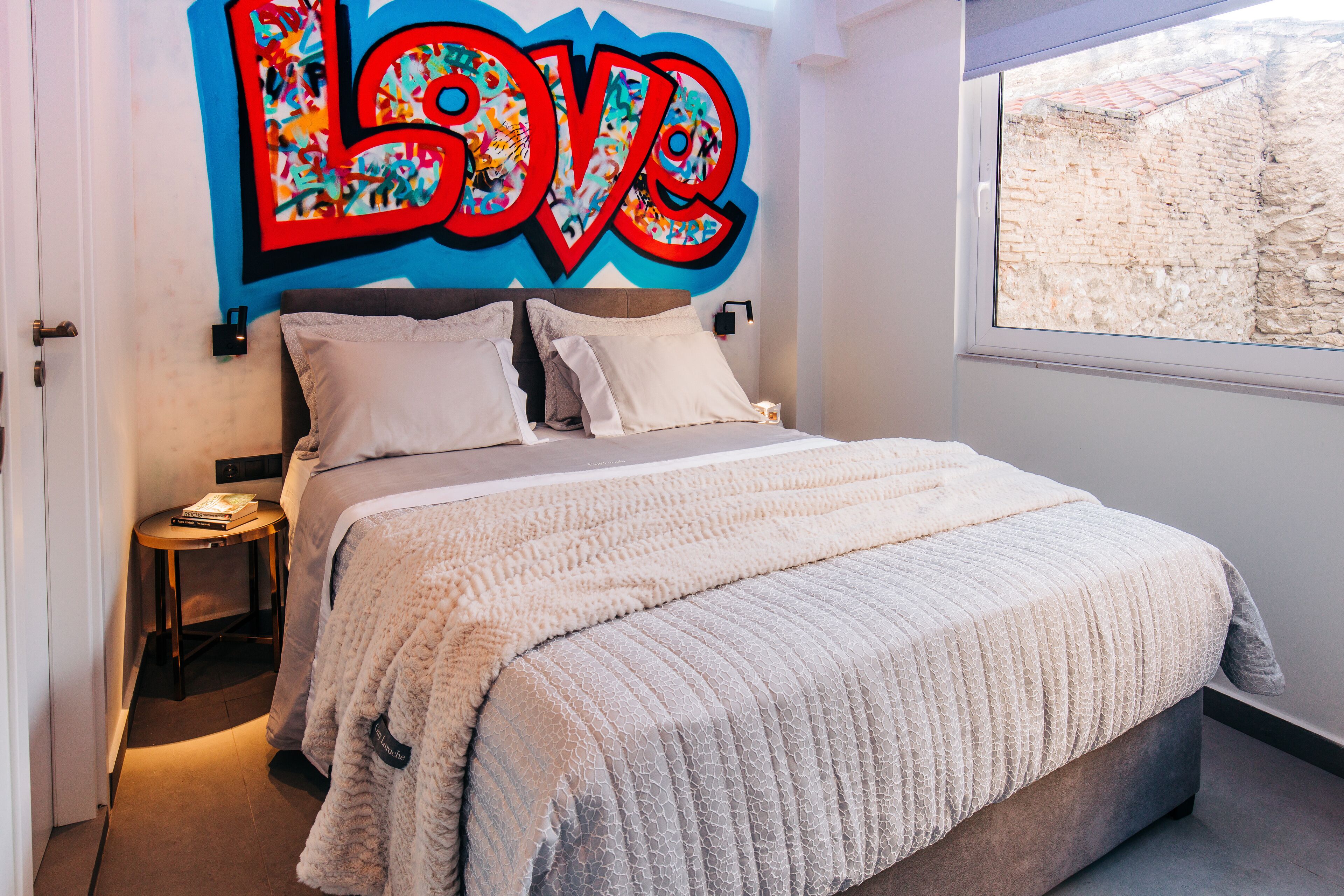 A bedroom with a large bed, a wall-mounted artwork with the word "LOVE," and a window with a view of a brick wall.