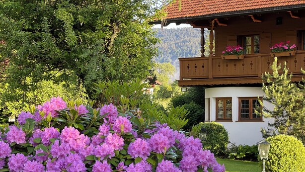 A traditional chalet with a red-tiled roof, surrounded by lush greenery and flowering plants. - Landhaus Christl am See (Bad Wiessee)
