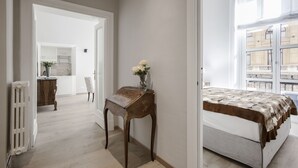 Ein Schlafzimmer mit einem Bett, einem Nachttisch mit Blumenvase und Blick auf Gebäude durch ein Fenster.