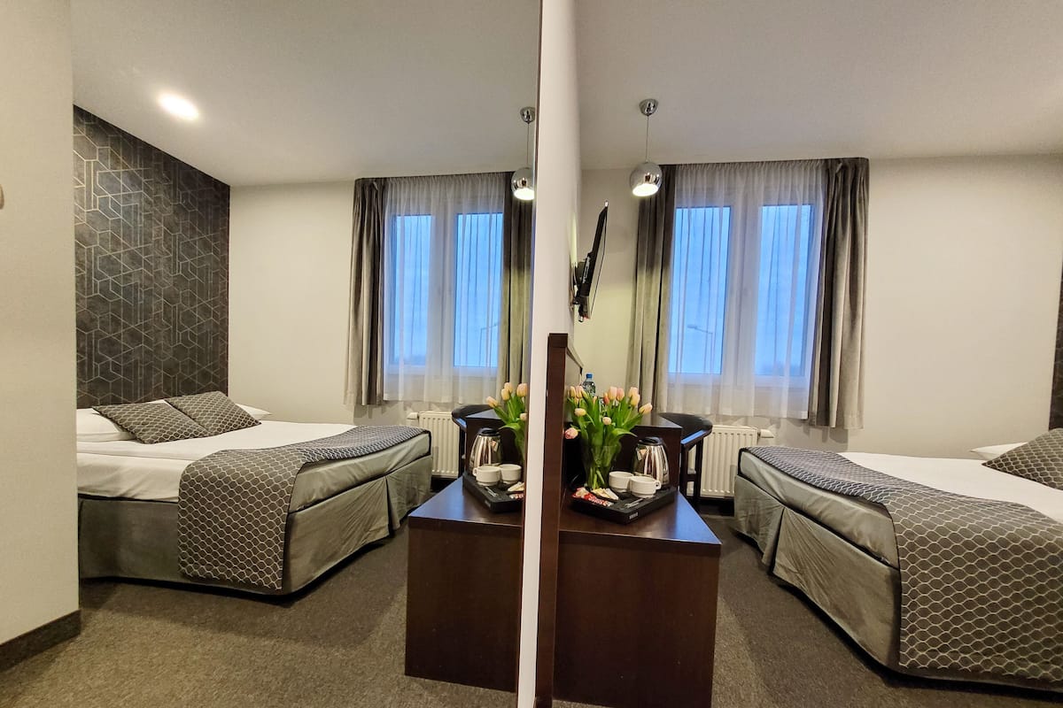 Quarto de hotel com duas camas, uma mesa de centro com um vaso de flores e vista da cidade por grandes janelas.