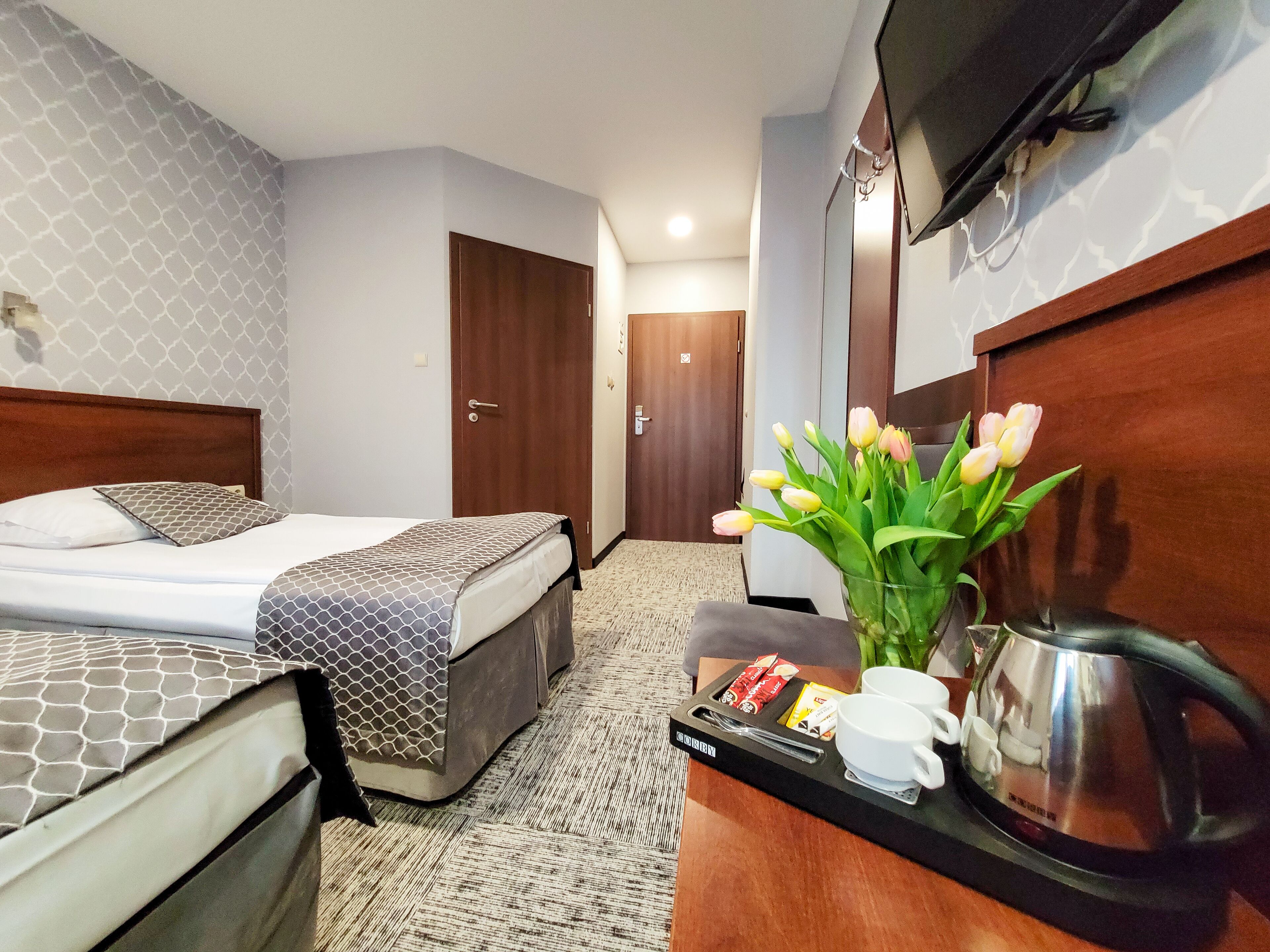 A hotel room with a bed, a desk with a kettle, a tray with cups and snacks, and a vase with flowers.