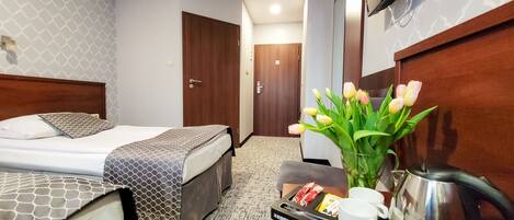 A hotel room with a bed, a desk with a kettle, a tray with cups and snacks, and a vase with flowers.