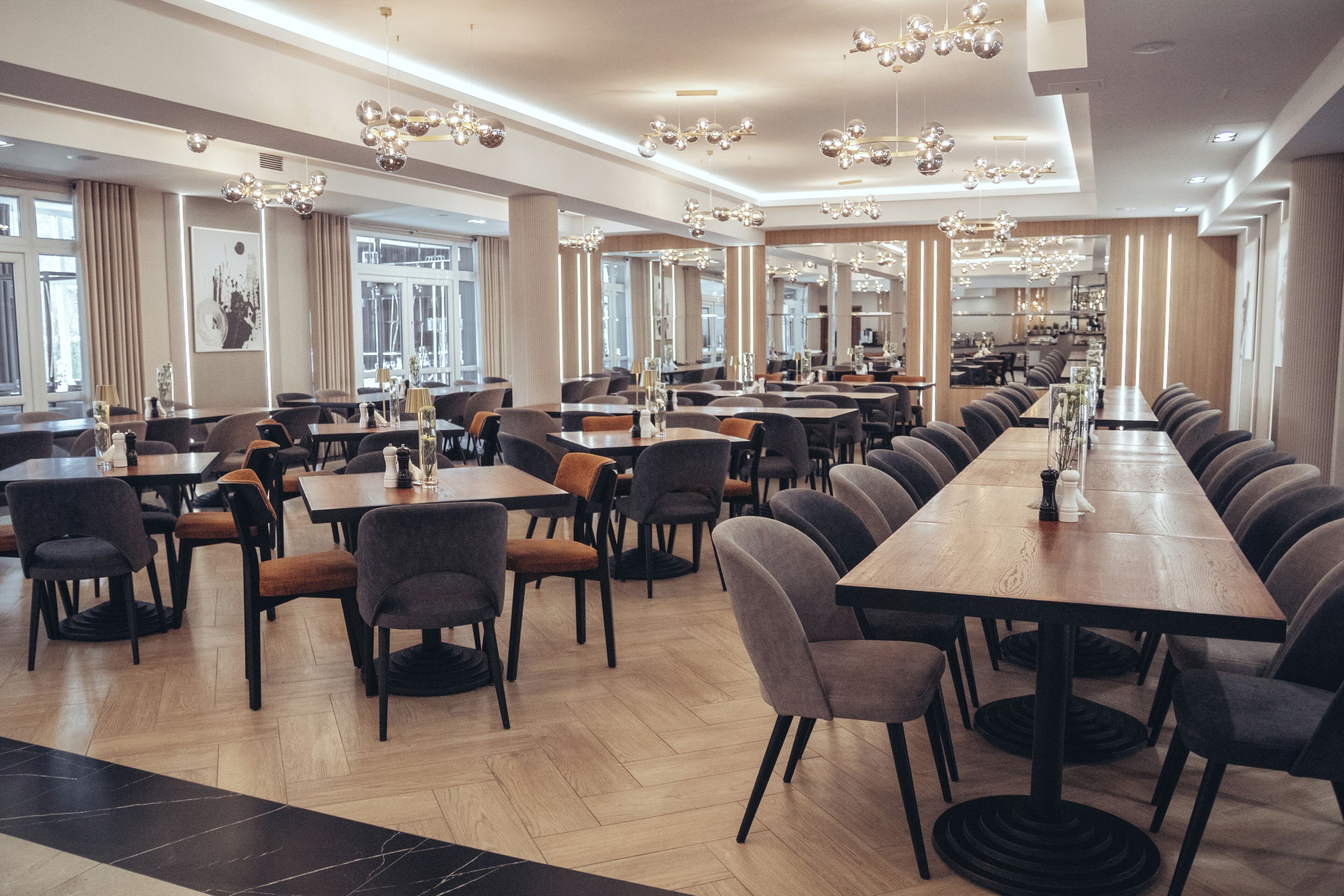 A spacious restaurant with multiple dining tables and chairs, large windows, and modern chandeliers.