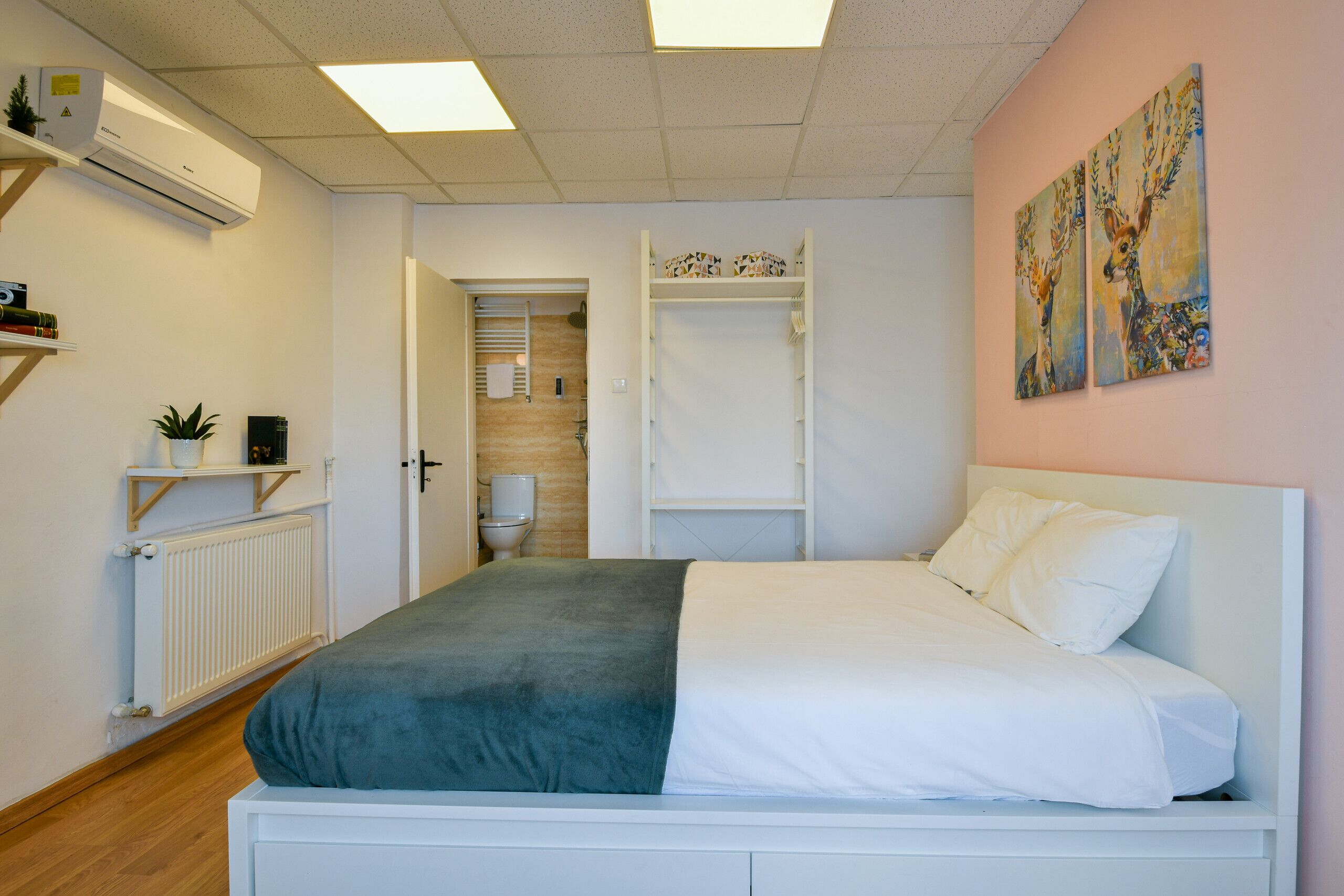 Ein Schlafzimmer mit Bett, Heizkörper, Ablagefläche mit Pflanze und einer Tür zu einem Badezimmer.