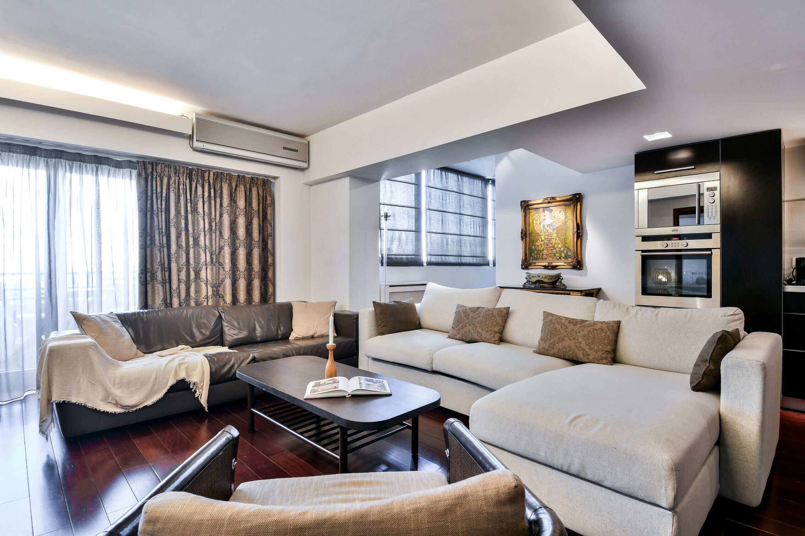 A modern living room with a sofa, armchair, and coffee table, an air conditioning unit, a painting, and a kitchen area with appliances.