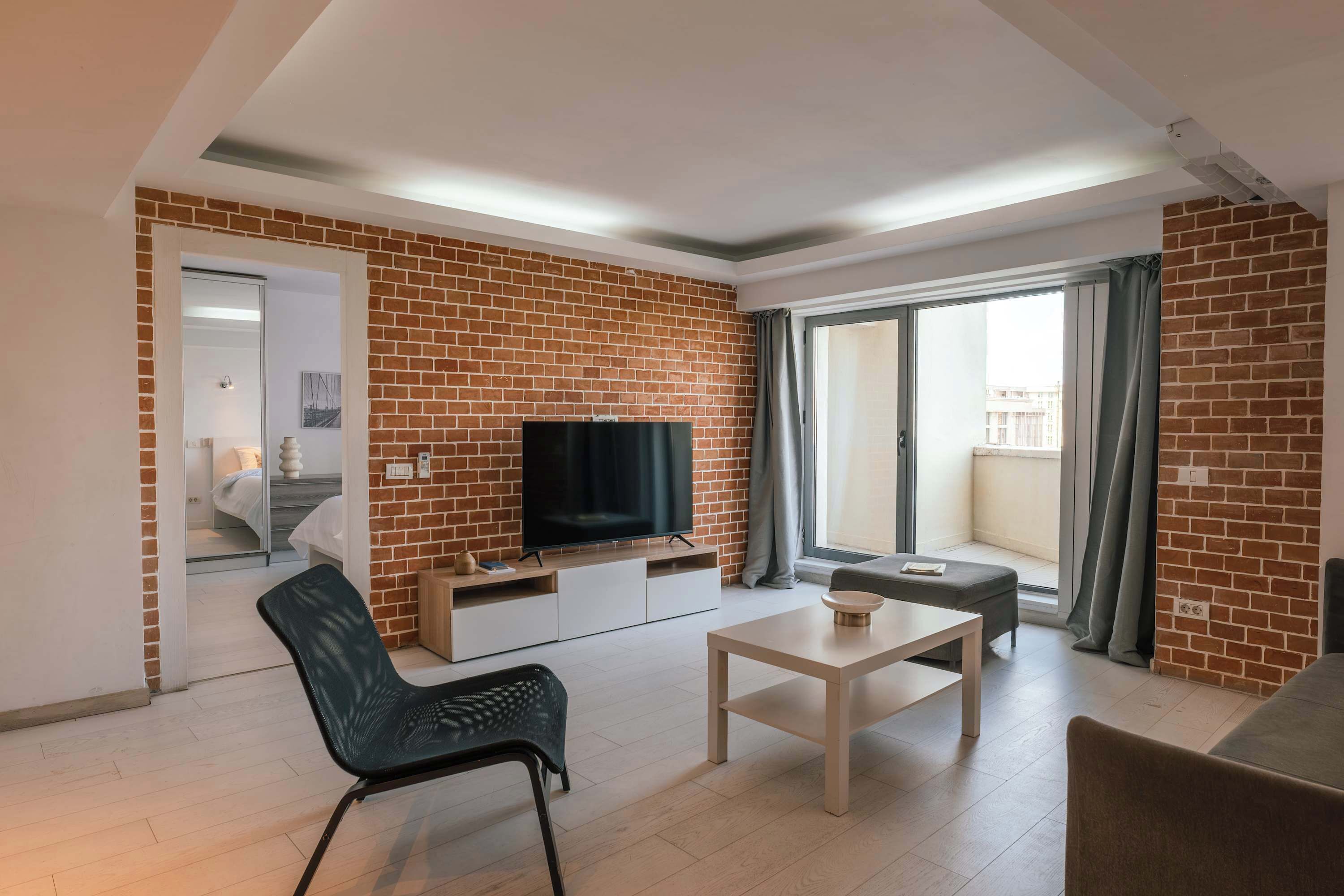 A modern living room with a brick wall, a flat-screen TV, a sofa, a coffee table, and a sliding glass door leading to a balcony.