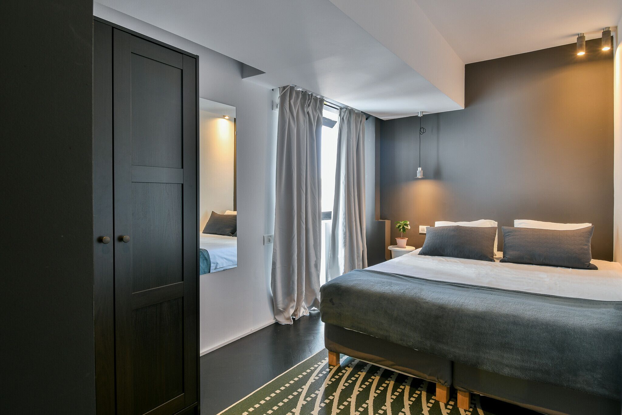 A bedroom with a bed, a wardrobe, a bedside table, a lamp, and a window with curtains.