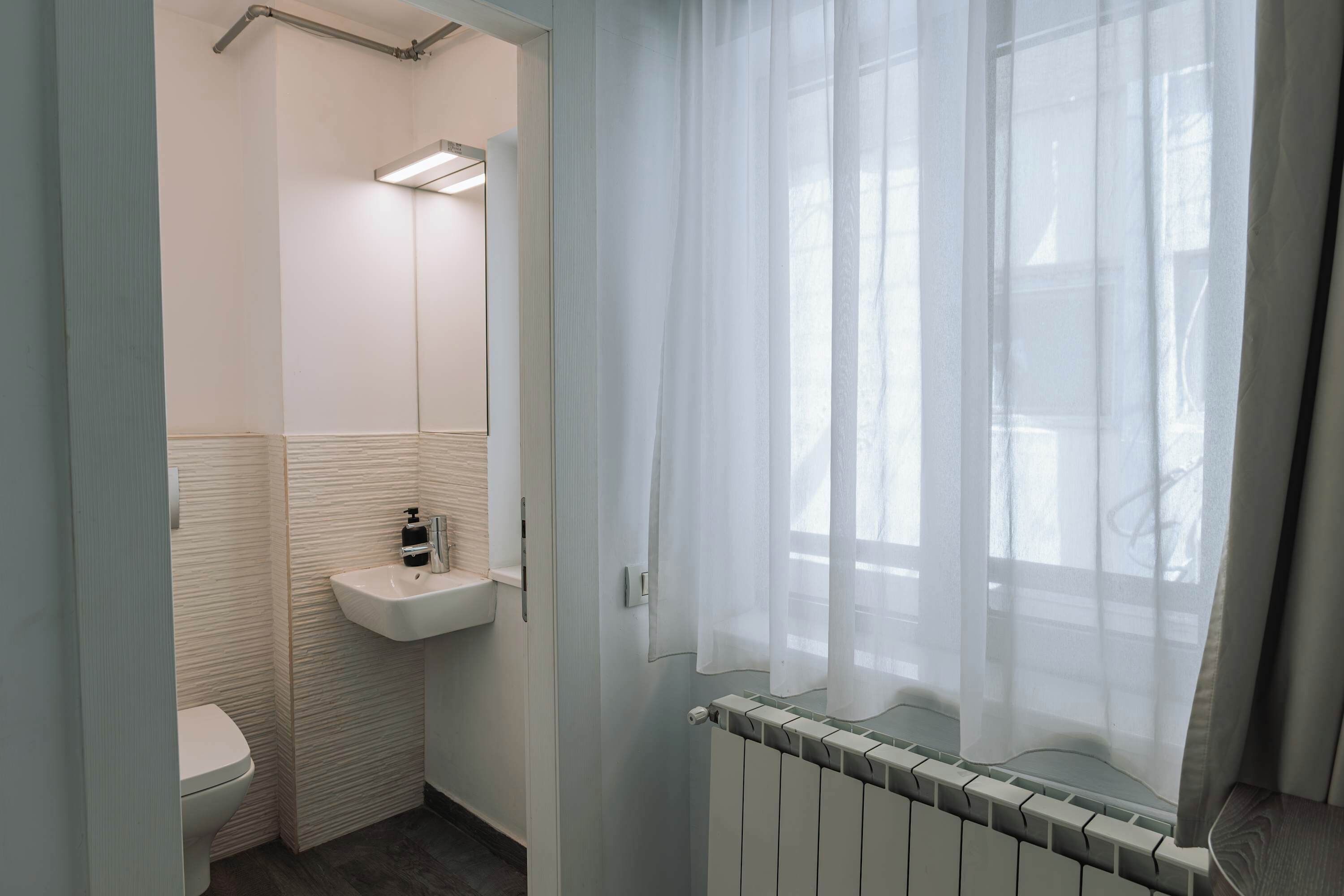 A bathroom with a toilet, sink, mirror, and a window with sheer curtains.
