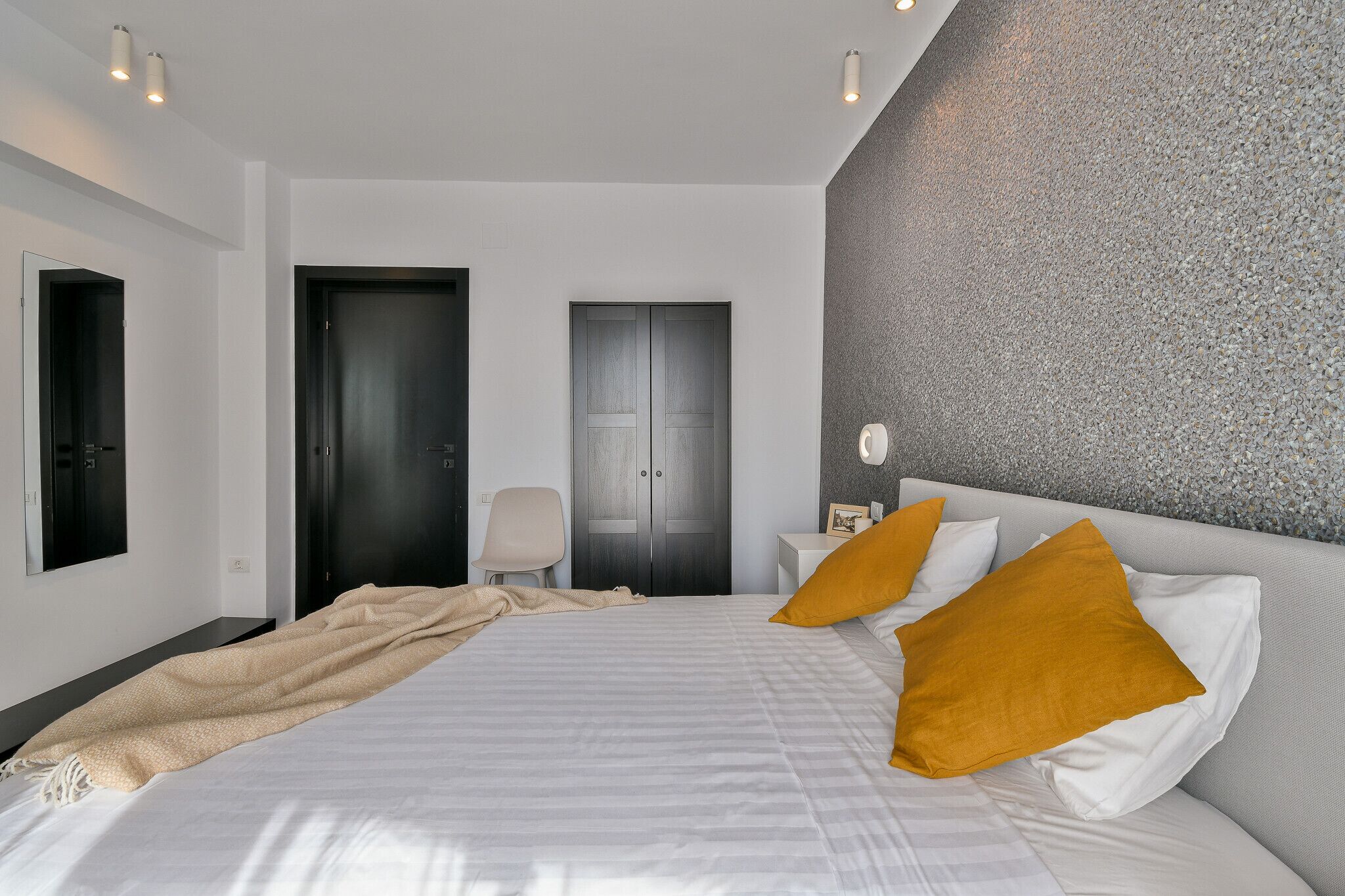 A modern hotel room with a large bed, a chair, a desk, and a textured wall.