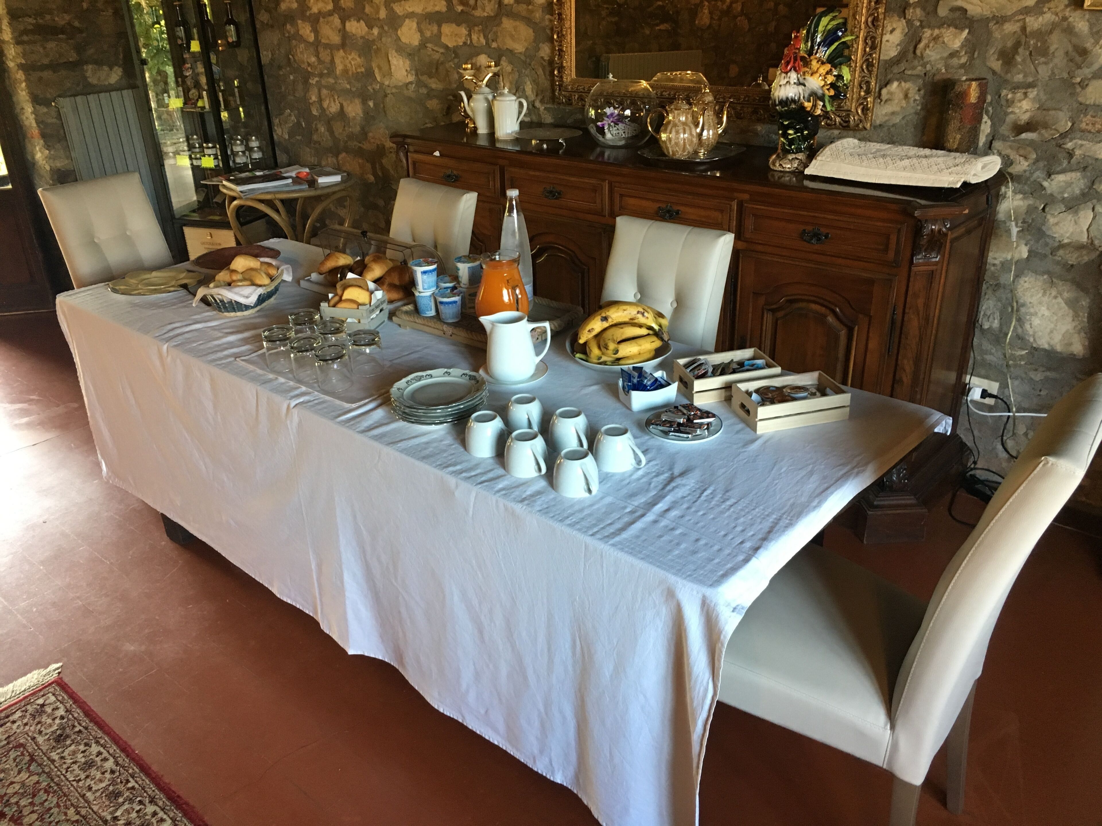 Una mesa de comedor dispuesta con diversos alimentos para el desayuno, como pan, pasteles y bebidas, en una habitación con paredes de piedra y decoración rústica.