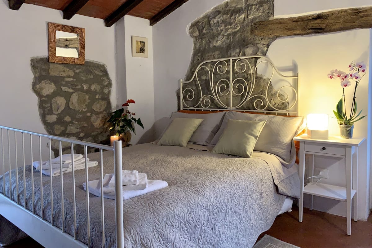 Un dormitorio con pared de piedra, cama con cabecera blanca y mesita de noche con lámpara y planta de orquídea.