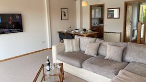 Uma sala de estar com sofá, mesa de centro de vidro e área de jantar com mesa posta para quatro pessoas.