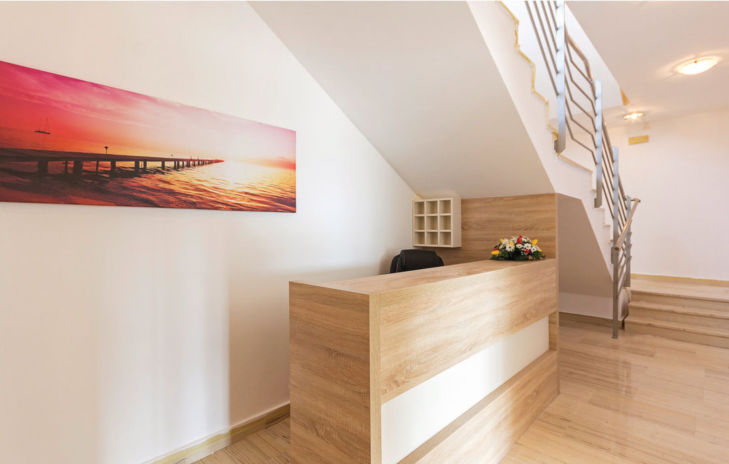 A reception desk with a wooden finish, a painting of a sunset over a pier, and a staircase with a metal railing.