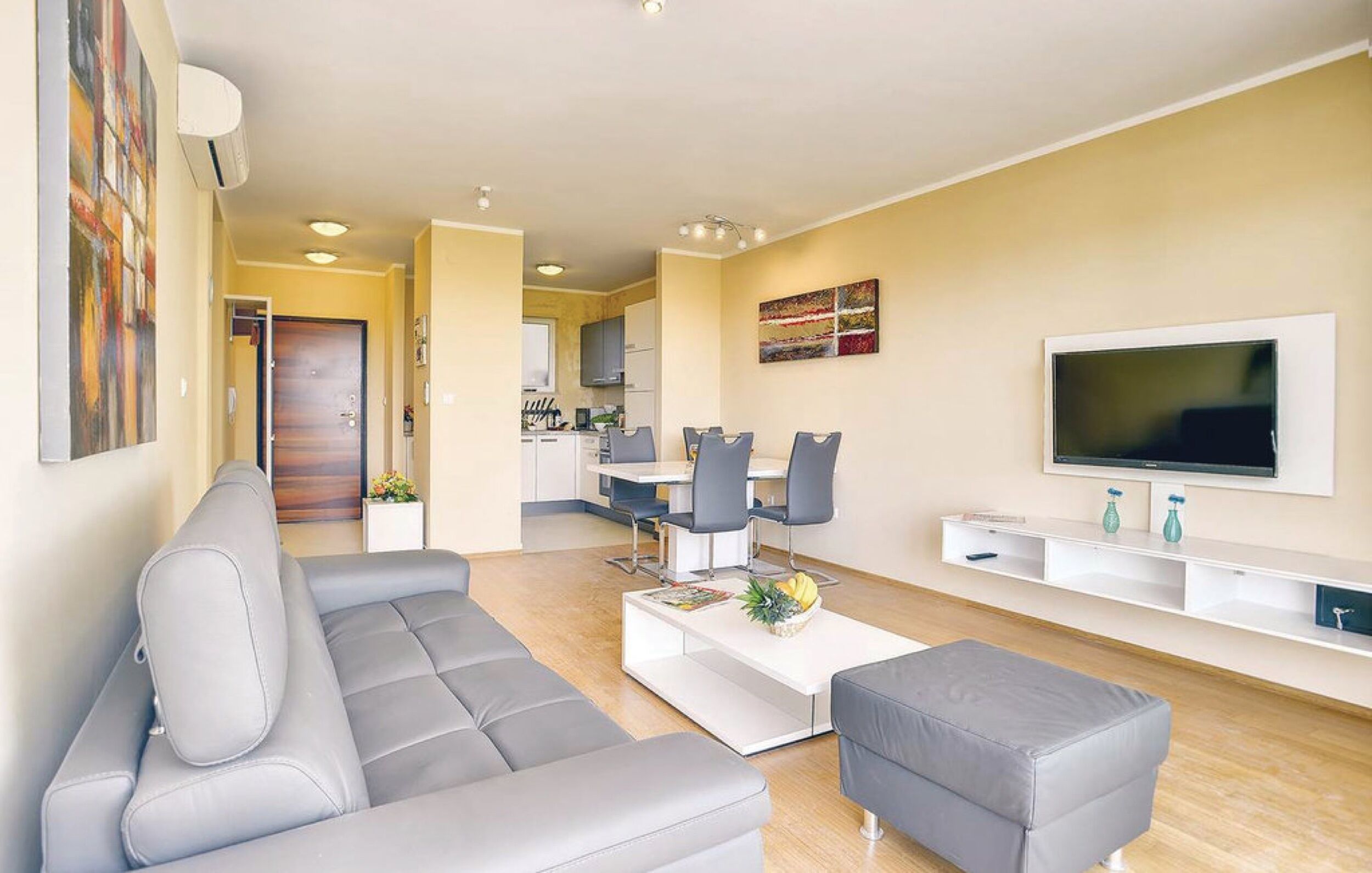 A modern living room with a grey sofa, a coffee table with fruits, a TV on the wall, and a kitchen in the background.