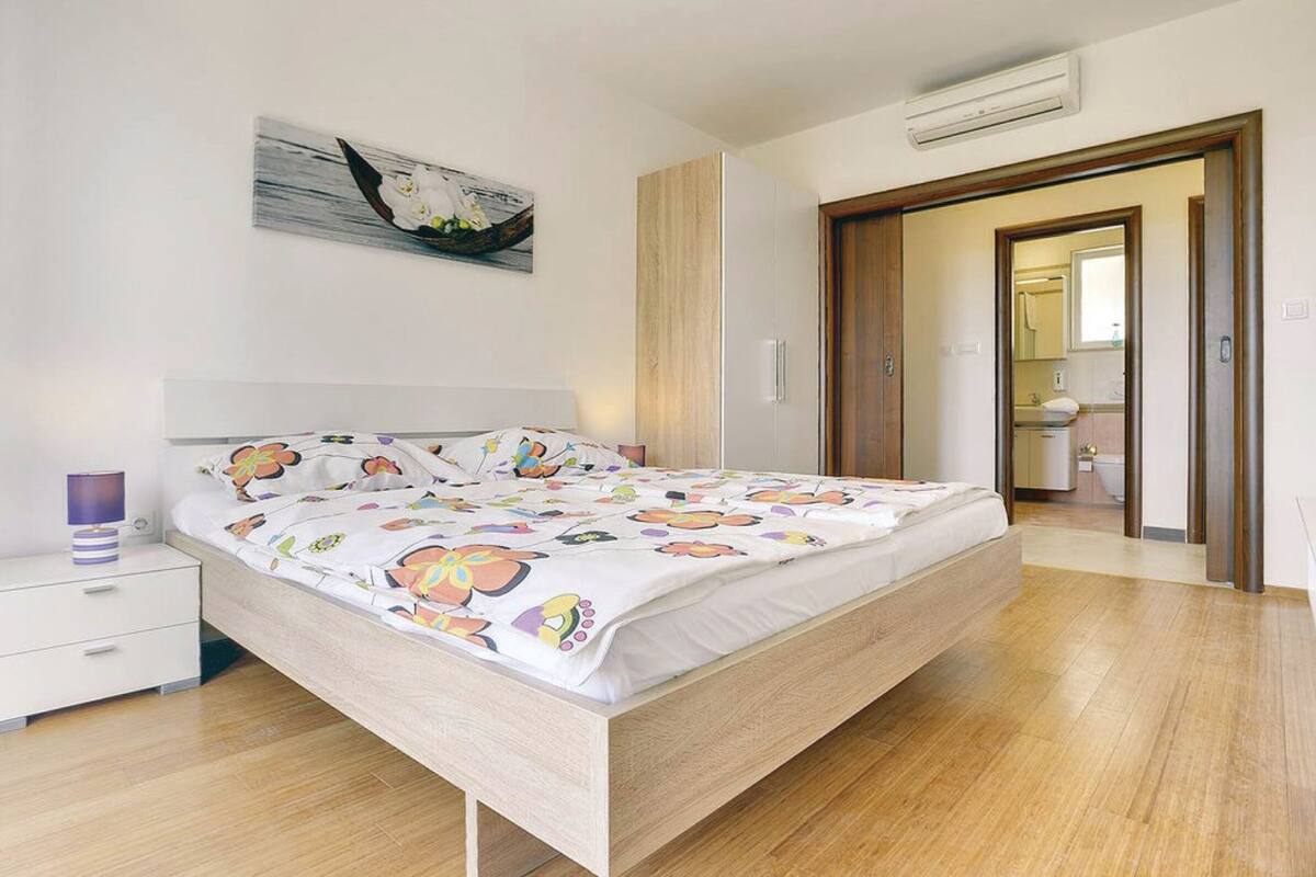 A bedroom with a wooden bed, a nightstand, a wall-mounted air conditioner, and a door leading to a bathroom.