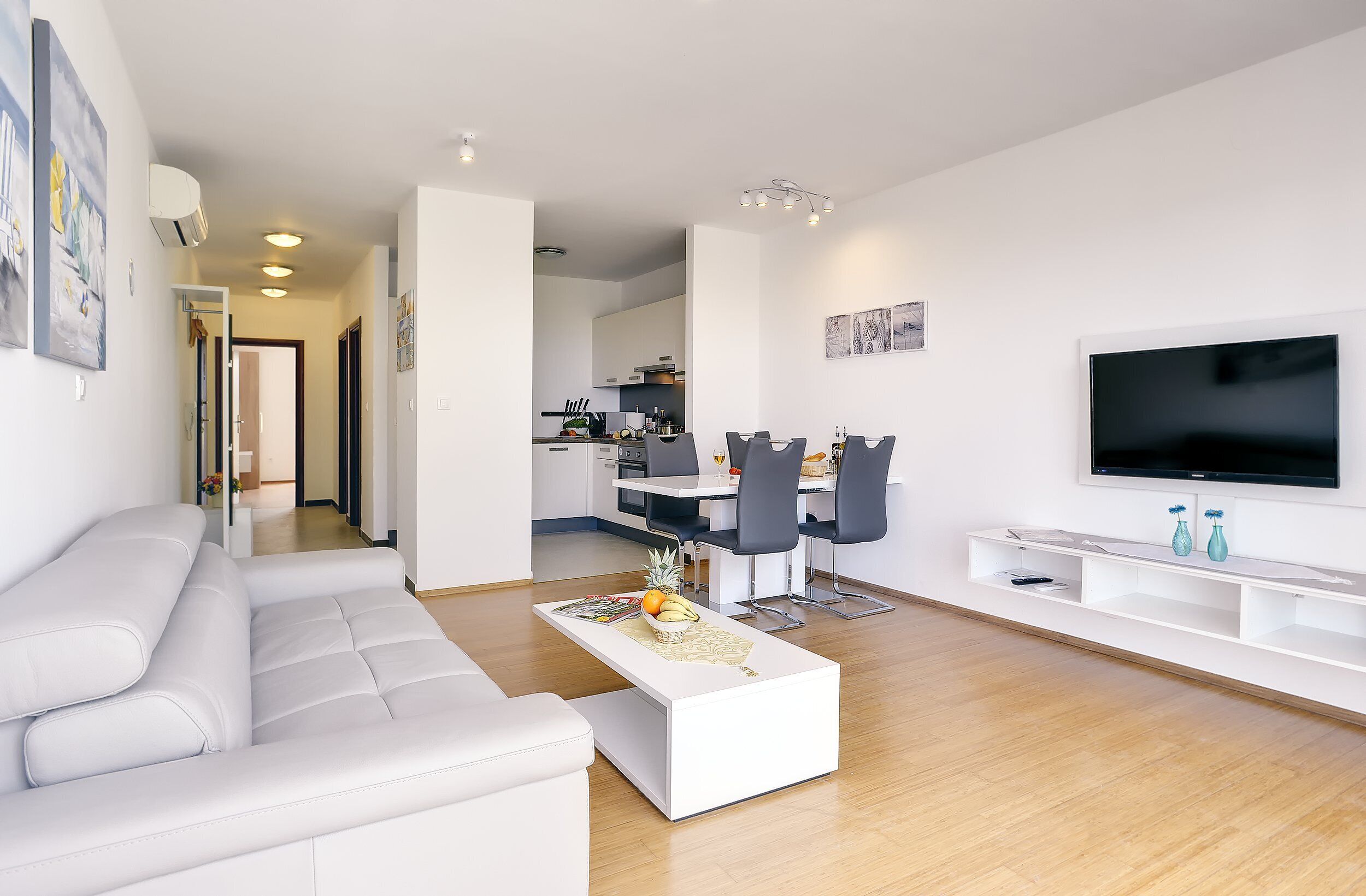 A modern living room with a white sofa, a coffee table with fruits, a dining area with chairs, and a TV on the wall.