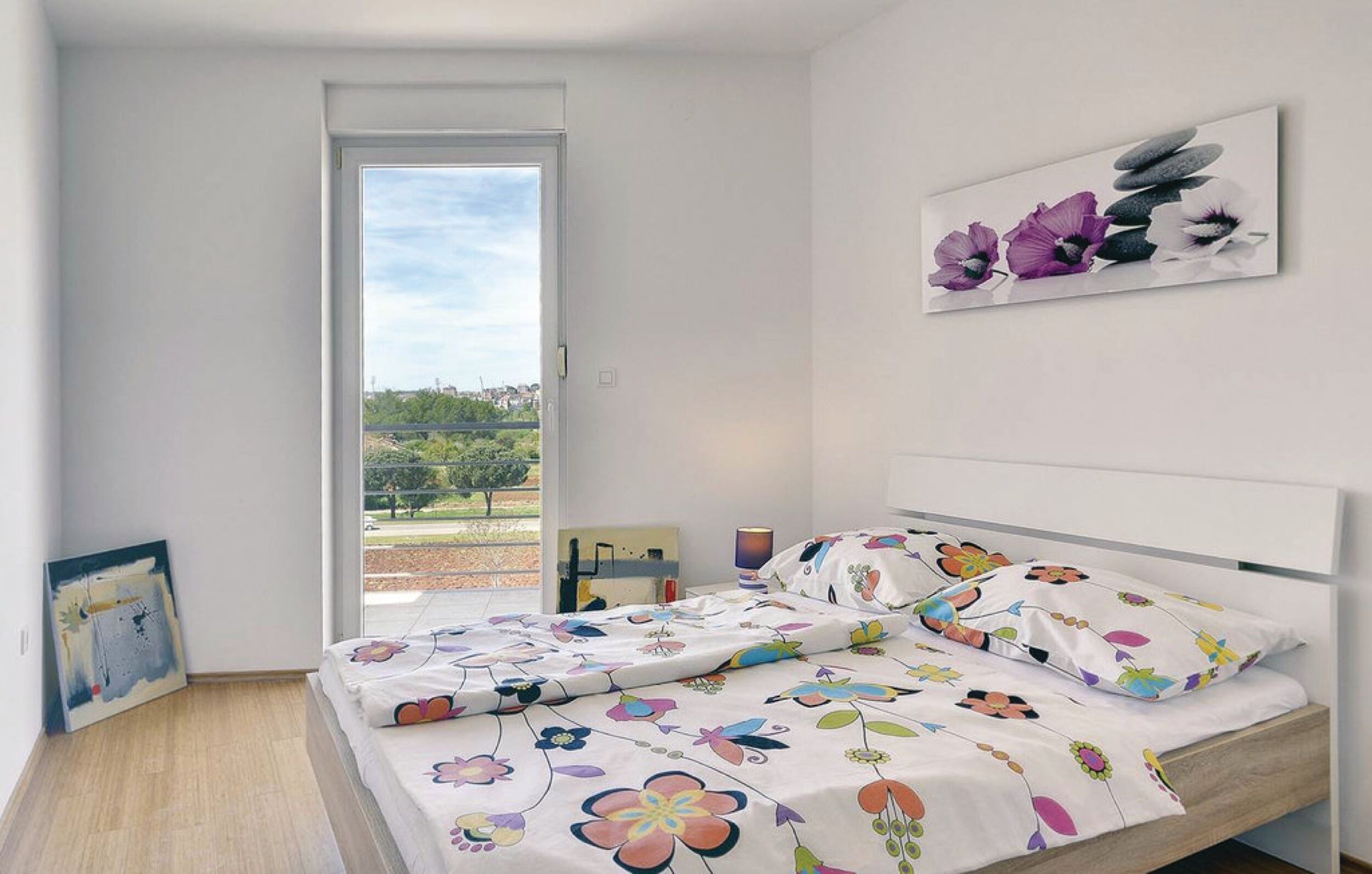 A bedroom with a bed, a painting on the wall, and a view of the outdoors.