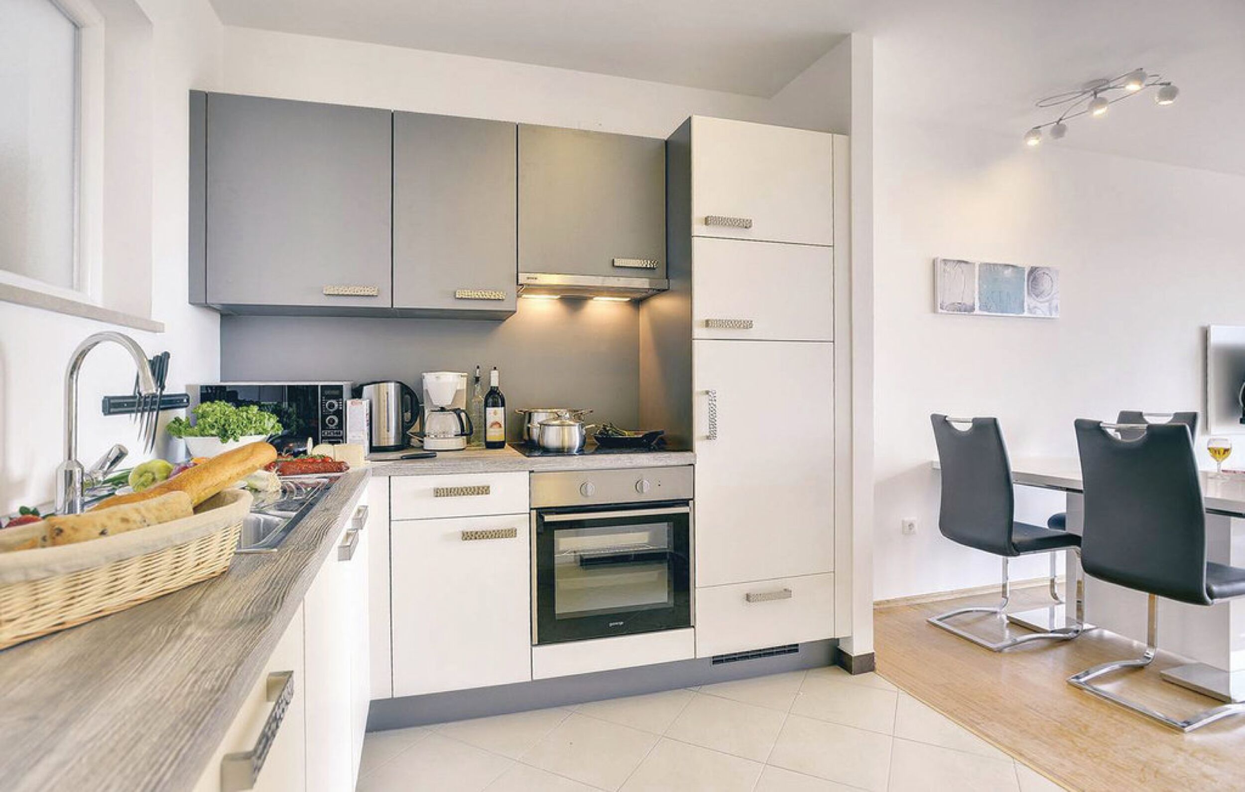 Una cocina moderna con armarios grises, un horno empotrado, una zona de comedor con dos sillas negras y una mesa pequeña.