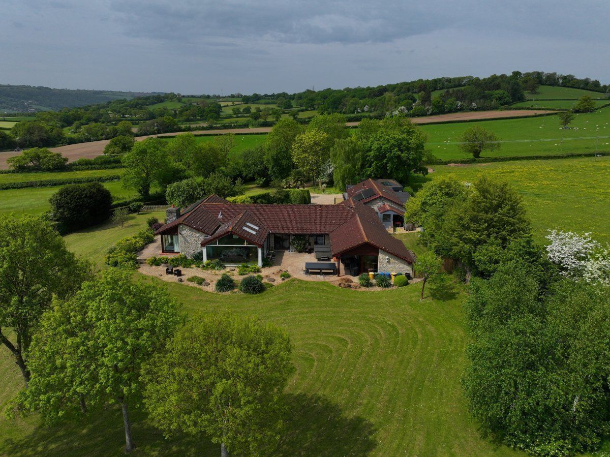 Une grande maison à la façade en pierre et au toit rouge, entourée de champs verdoyants et d’arbres.