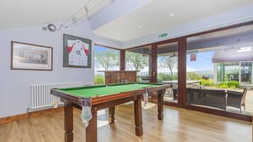 Uma sala de bilhar com mesa de sinuca, quadros nas paredes e vista para o exterior por grandes portas de vidro.