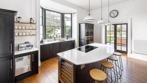 A modern kitchen with a central island, bar stools, and a view of the outdoors through large windows. - Cotswold Manor House - Group Accommodation in The Cotswolds (Badminton)