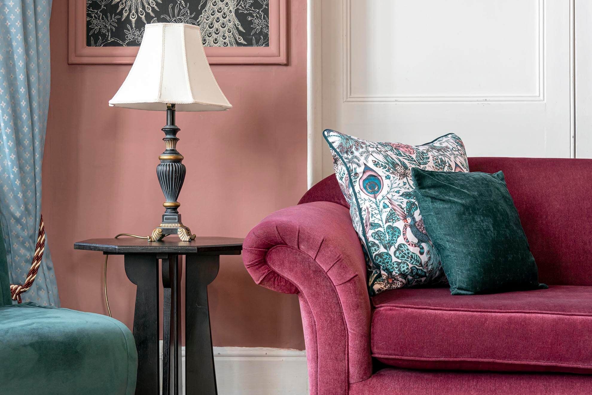 A room with a red sofa, a patterned pillow, a lamp on a small table, and a curtain with a floral pattern.