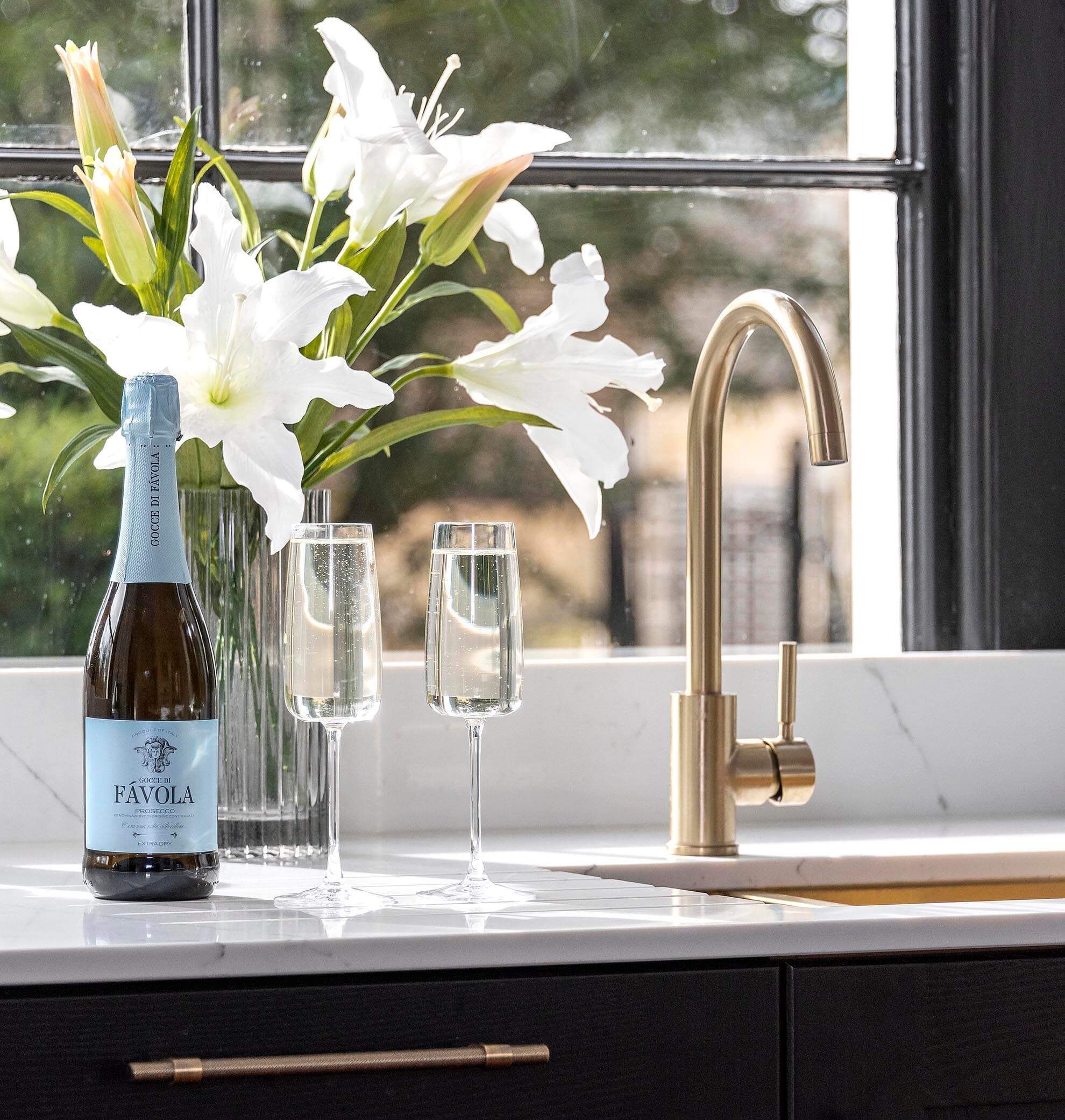 A bottle of Favola Prosecco, two glasses, and a gold faucet on a kitchen counter by a window.