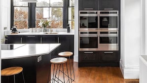 A modern kitchen with a central island, black cabinetry, stainless steel appliances, and wooden flooring. - Cotswold Manor House - Group Accommodation in The Cotswolds (Badminton)