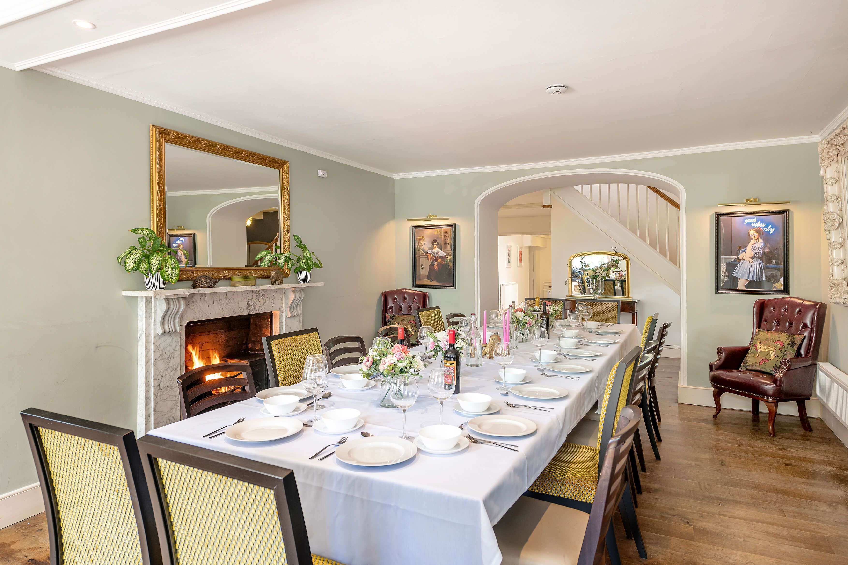 A dining room with a long table set for a meal, a fireplace, and a staircase leading to another level.