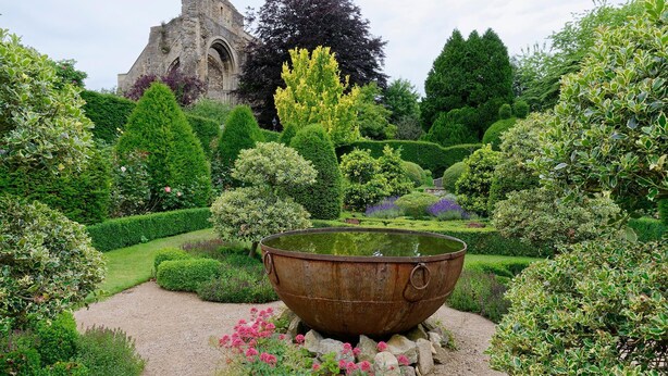 A garden with a large, rusted metal basin in the center, surrounded by well-manicured hedges and flowering plants. In the background, there is a stone structure with arches. - Cotswold Manor House - Group Accommodation in The Cotswolds (Badminton)