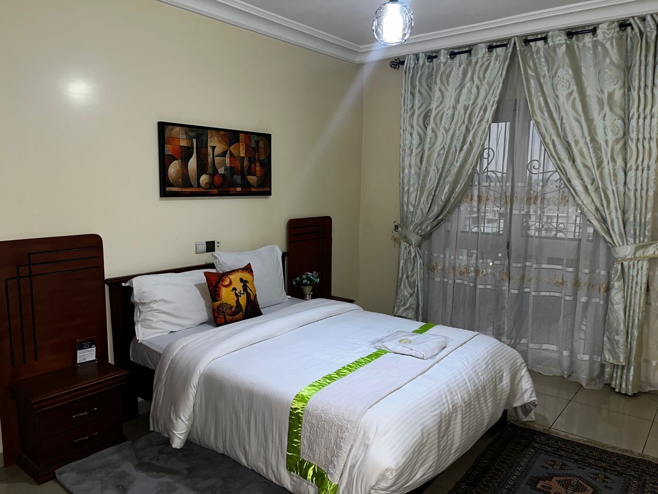 Una cama bien hecha con detalles en verde, una mesita de noche, un cuadro colgado en la pared y una ventana con cortinas translúcidas.