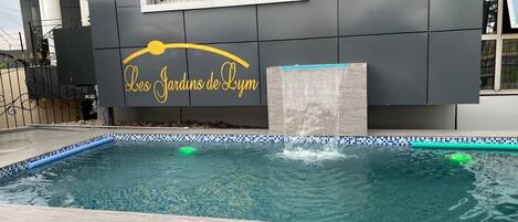 Ein Schwimmbecken mit einer gefliesten Kante, einem Wasserspiel und einem Gebäude mit der Aufschrift 'Les Jardins de Lym'.