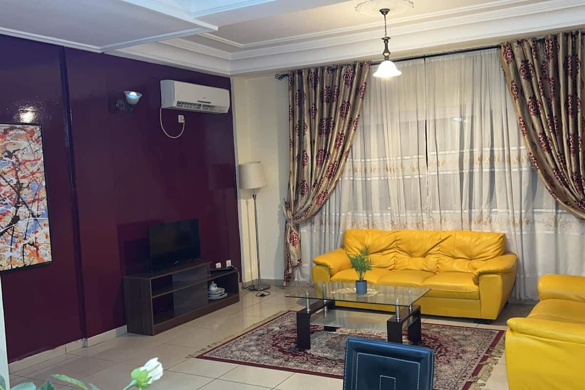 Una sala de estar con un sofá amarillo, una mesa de centro de vidrio, un televisor de pantalla plana y un equipo de aire acondicionado.