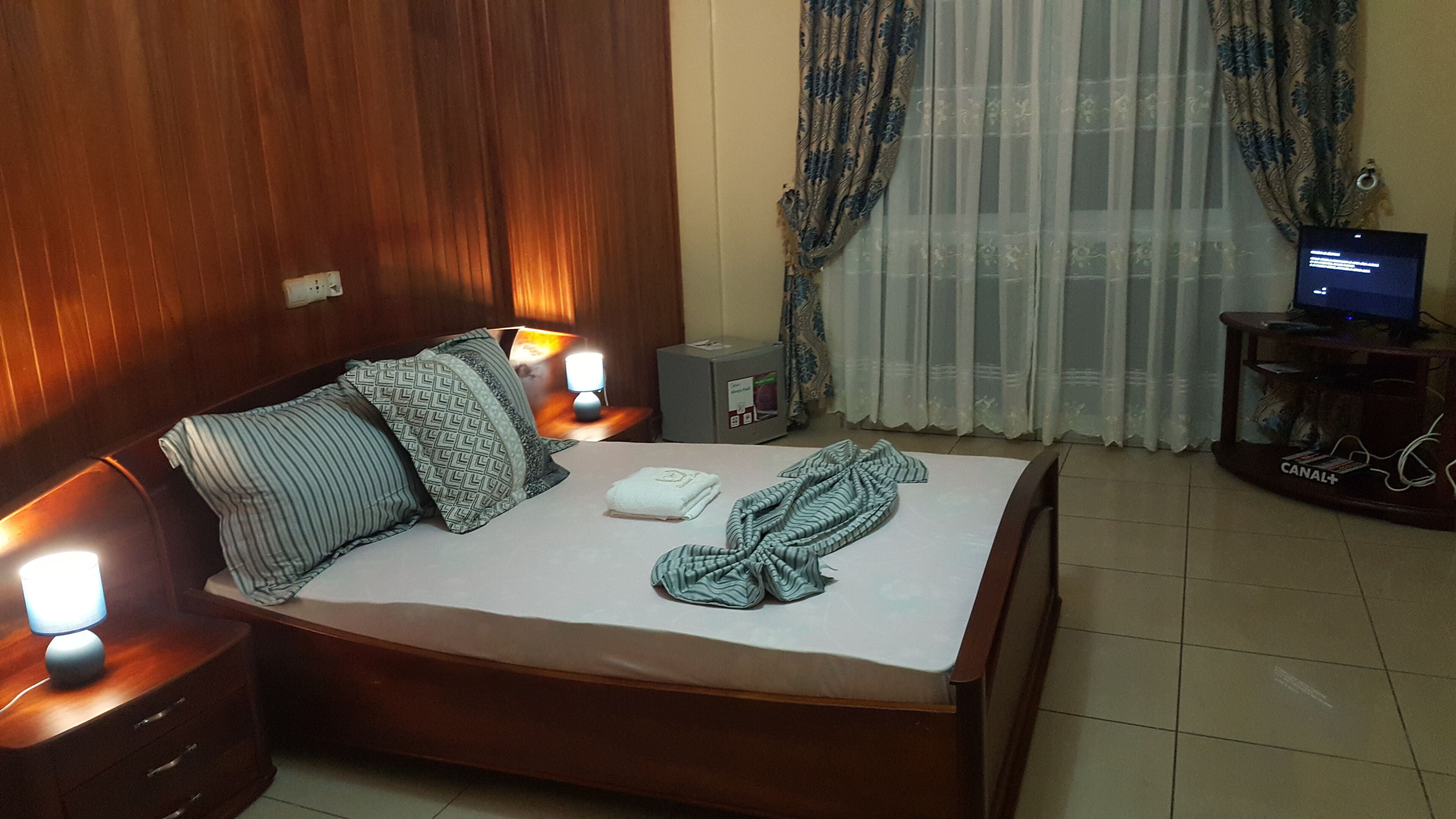 A hotel room with a bed, pillows, a TV, a small cabinet, and a lamp.