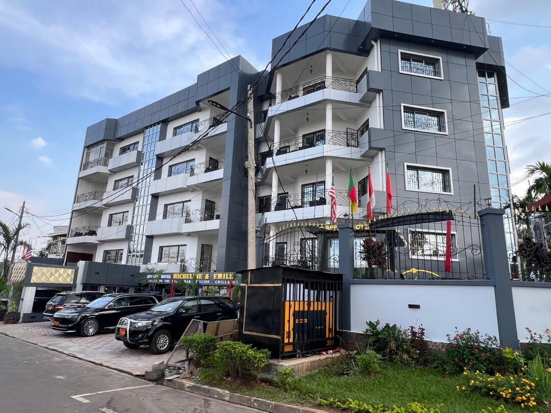 Hotel com vários andares, varandas, portão fechado e estacionamento com carros.