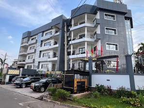 Un hotel de varios pisos con balcones, una entrada con reja y un estacionamiento con coches.