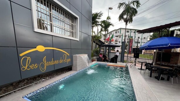 A swimming pool with a tiled edge, a covered seating area, and a sign reading 'Les Jardins de L'Hotel.' - Résidences hôtelières Micheline et Emile, Bastos (Yaoundé)