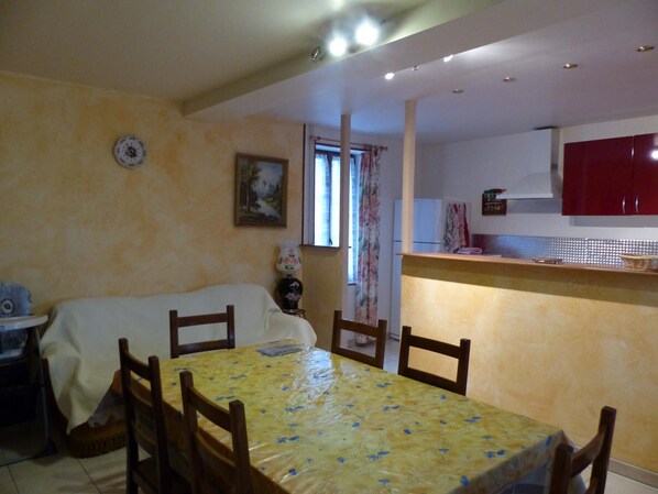 Un coin repas avec une table recouverte d’une nappe jaune, des chaises en bois, un canapé, une petite table avec une lampe, et un espace cuisine avec des placards rouges.