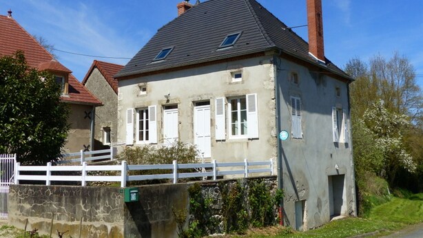 Une maison à deux étages, avec une façade grise, des volets blancs et un toit recouvert de tuiles rouges.
