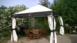 Un belvédère avec une table et des chaises, entouré de verdure et d’un arbuste à fleurs blanches.