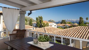 A terrace with a wooden table and chairs, a glass vase with plants, and a view of coastal houses and cliffs. - Villetta has two levels with beautiful sea view and located in front of a small park for children an (Golfo Aranci)
