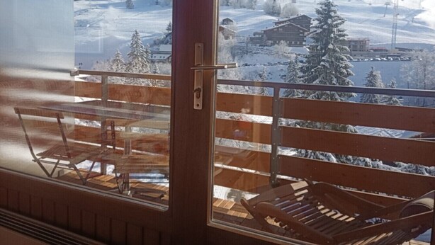 A wooden balcony with a view of a snowy mountain landscape, including a ski lift and buildings. - Appartement Plein Centre du Chinaillon (Le Grand-Bornand)