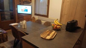 A kitchen with a wooden table, a fruit bowl, a cutting board, a coffee maker, and a television displaying a show. - Appartement Plein Centre du Chinaillon (Le Grand-Bornand)