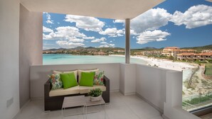 A balcony with a sofa and coffee table, overlooking a beach and resort area. - AIRONI - AIRONI 8 MONO (Golfo Aranci)