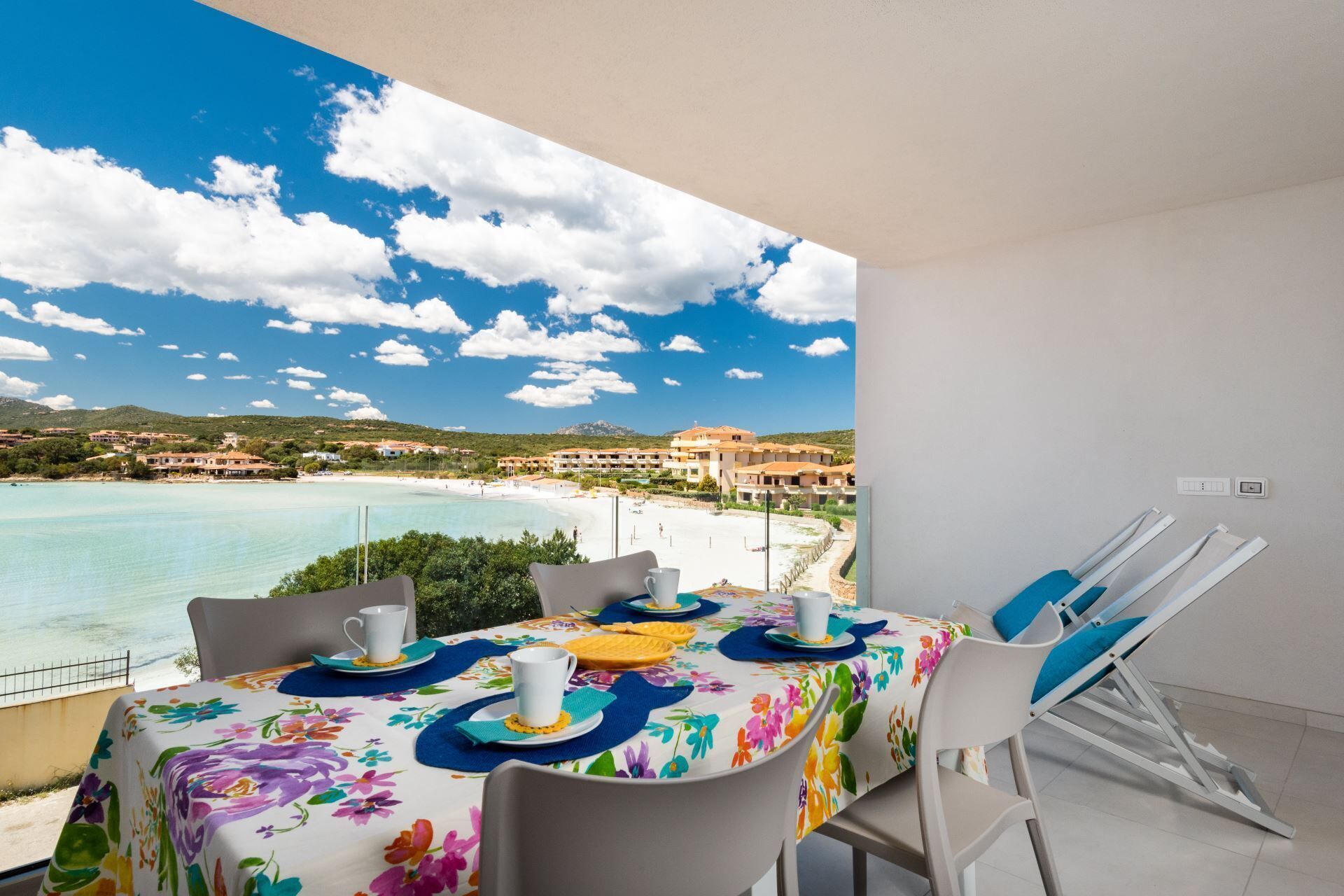 Un balcón con una mesa lista para dos personas, con vista a una playa y un pueblo costero.