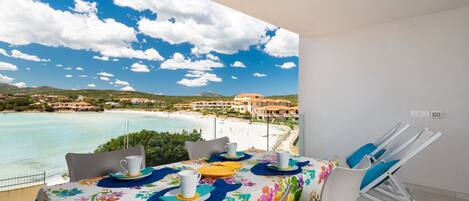 Un balcón con una mesa lista para dos personas, con vista a una playa y un pueblo costero.