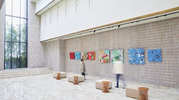 An interior with a high ceiling, a large window, and a wall with abstract art. There are seating areas with stools and a bench.